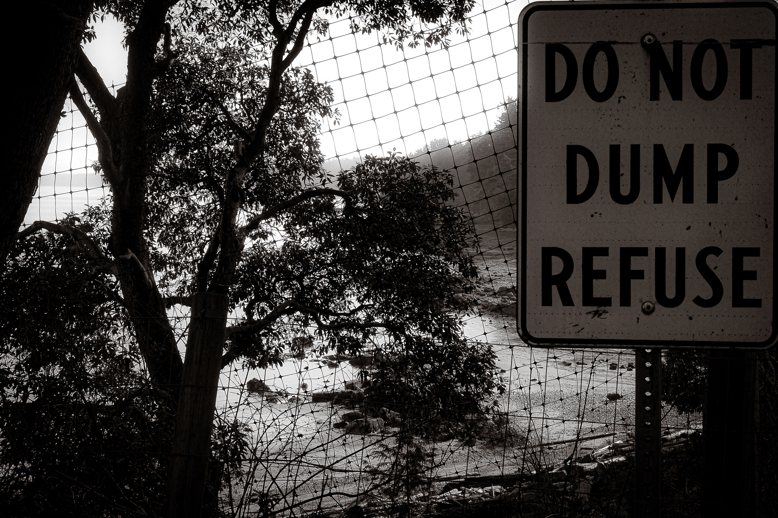 A refuse warning sign is seen through a wire fence beside a tree.