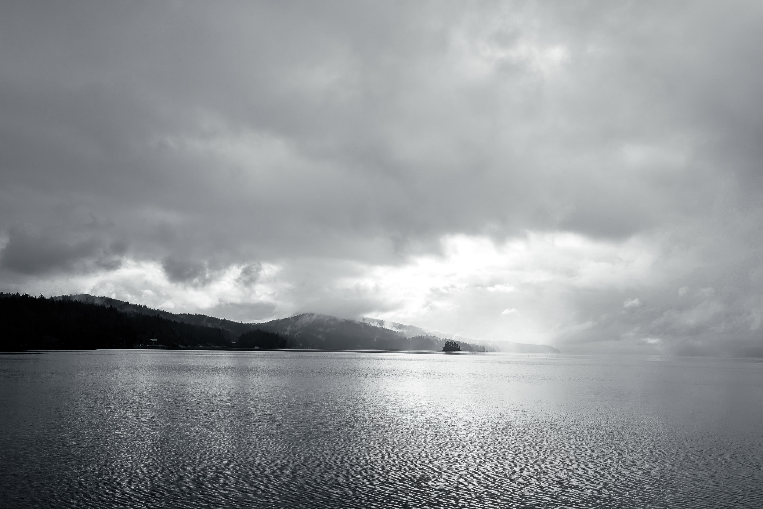 A dense bank of clouds moves over water and a low mountainous shore.